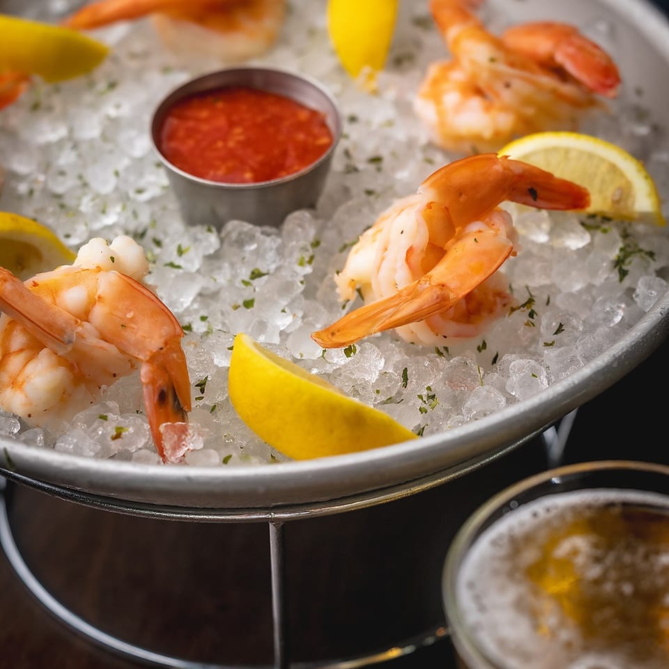 Chilled shrimp platter served on crushed ice with lemon wedges, cocktail sauce, and a sparkling drink.