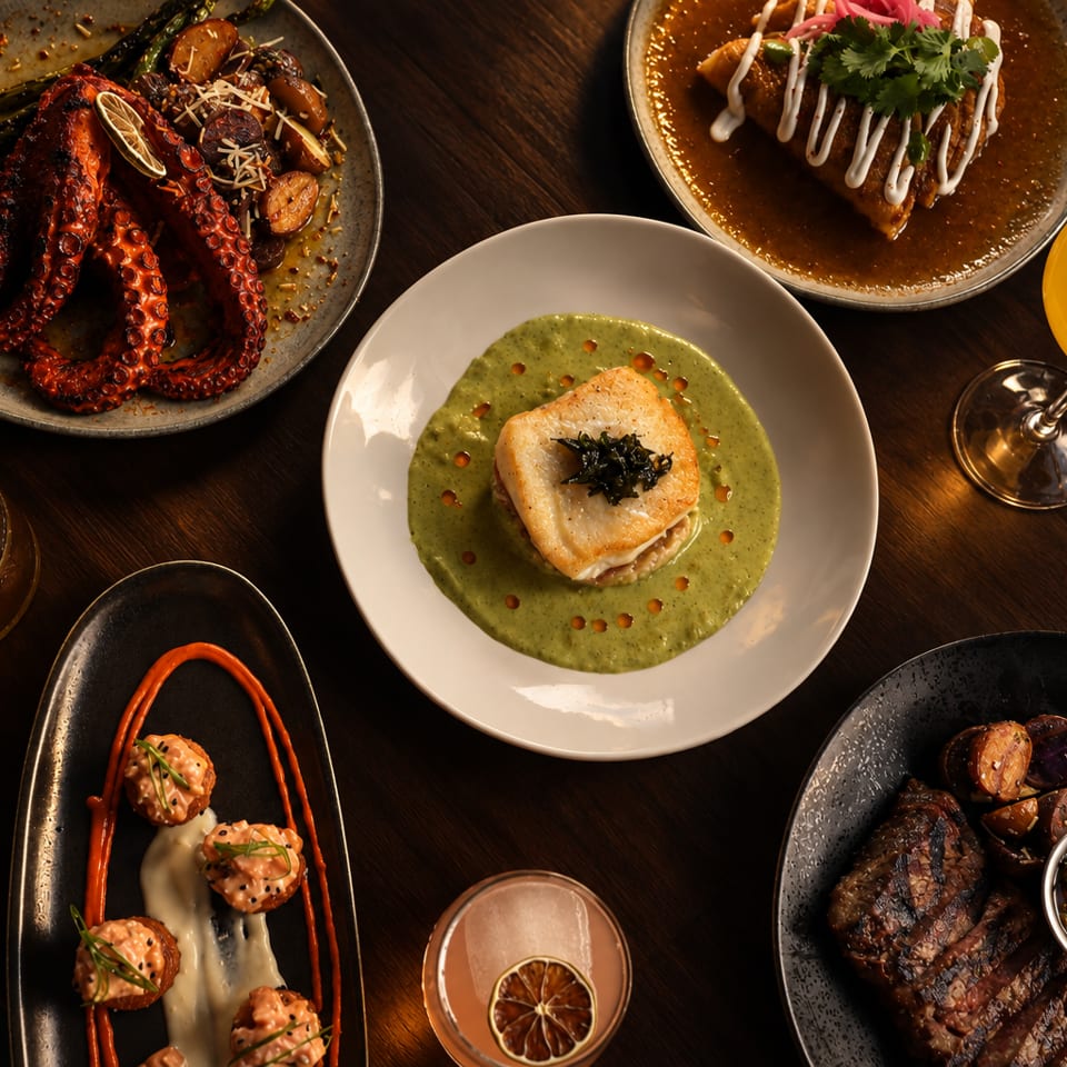 Overhead view of an upscale spread featuring grilled octopus, seared fish, sliced steak, garnished appetizers, and colorful cocktails on a dark wood table.