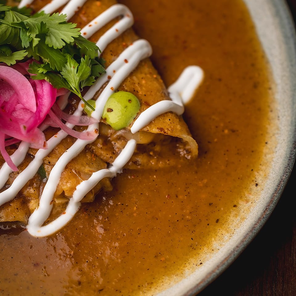 Enchiladas covered in brown sauce and cream, garnished with cilantro, pickled onions, and a lime wedge.