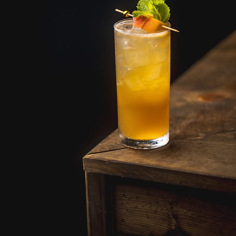 Golden iced cocktail garnished with fresh mint and peach wedges, served in a tall glass on a rustic wooden table.