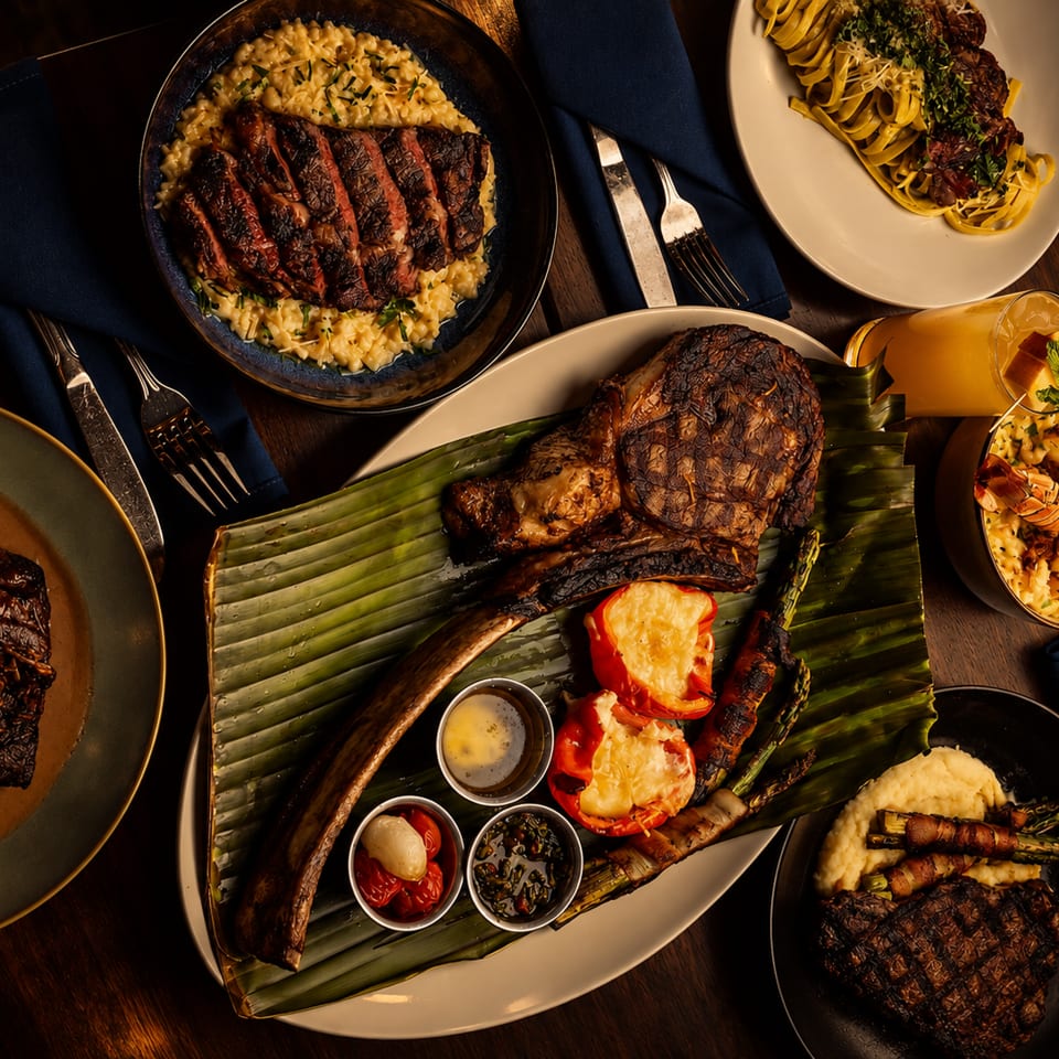Overhead spread of grilled steaks, lobster, shrimp, pasta, mashed potatoes, and vegetables on a warmly lit wooden table.