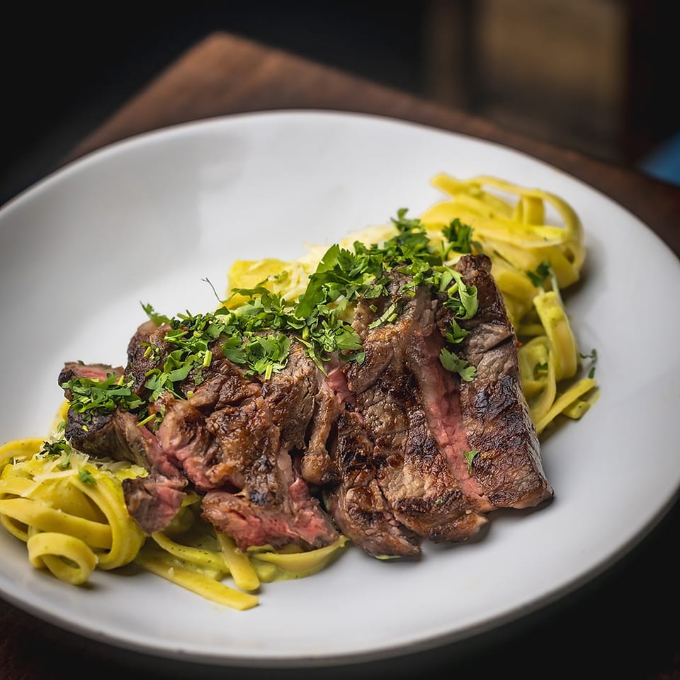 Grilled medium-rare steak topped with fresh herbs, served over yellow pasta on a white plate.
