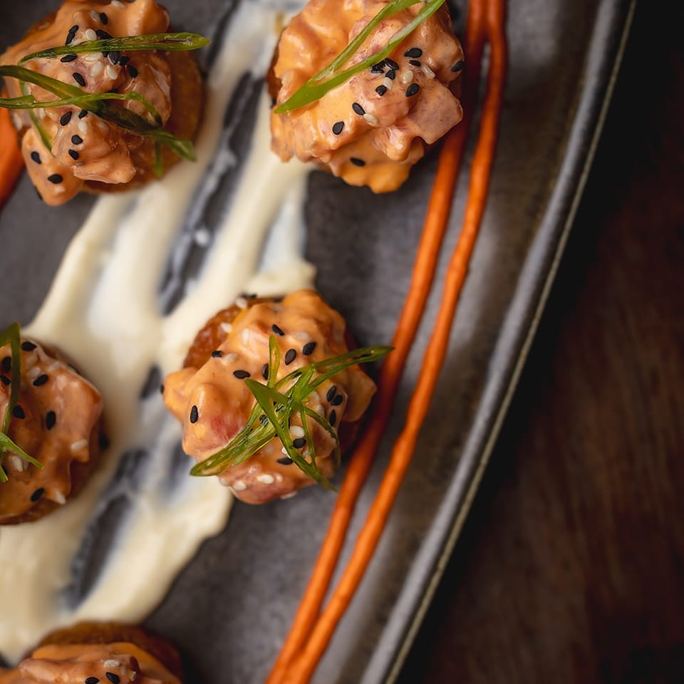 Glazed orange-brown bites topped with sesame seeds and scallions, arranged on a dark plate with cream and orange sauces.