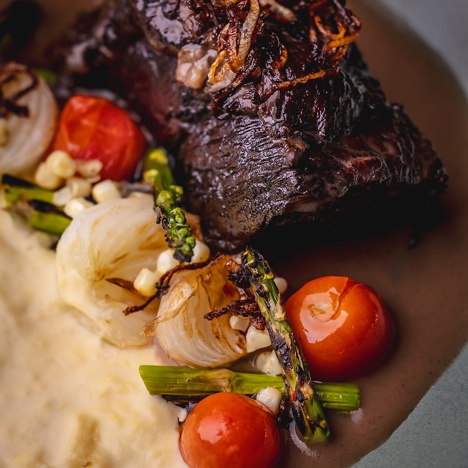 Seared steak topped with crispy onions, served with mashed potatoes, roasted tomatoes, onions, corn, and asparagus.