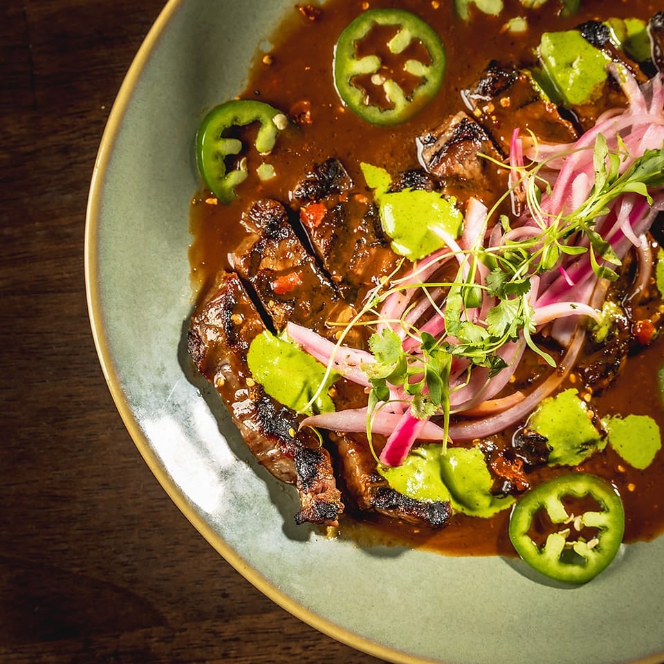 Grilled steak slices in a rich sauce, topped with pickled red onions, jalapeños, and green crema.