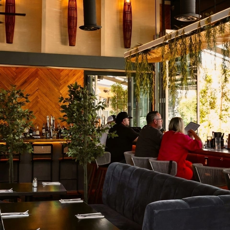 Restaurant interior with patrons at a bar, lush greenery, and modern decor with wooden accents.