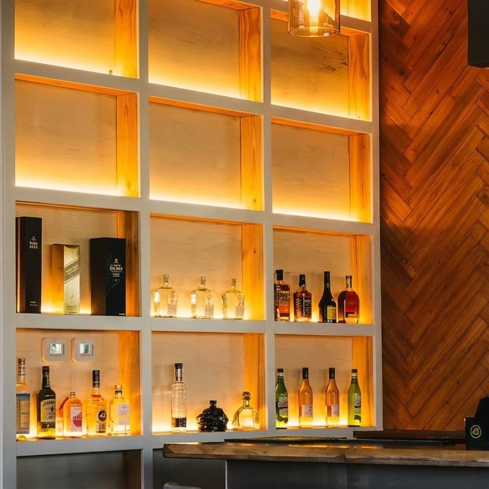 Backlit bar shelves display various liquor bottles, next to a wooden herringbone wall and rope curtain.